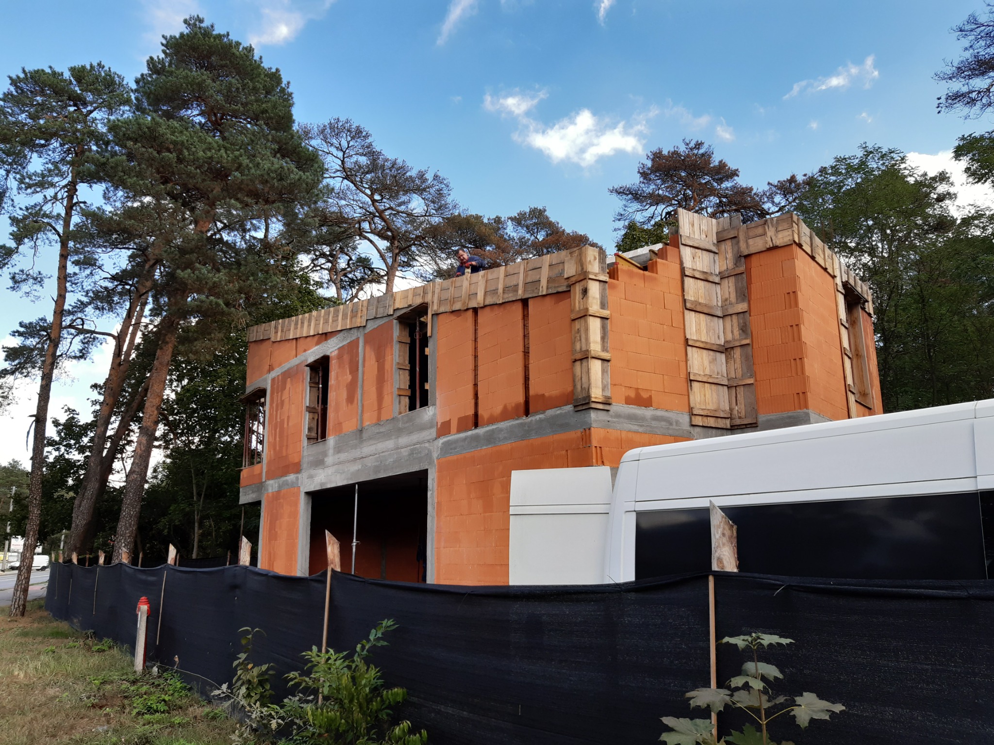 Wznoszenie dwukondygnacyjnego domu z czerwonej cegły, widok z poziomu gruntu, częściowo zasłonięty czarną siatką. Widoczny pracownik na górze konstrukcji.