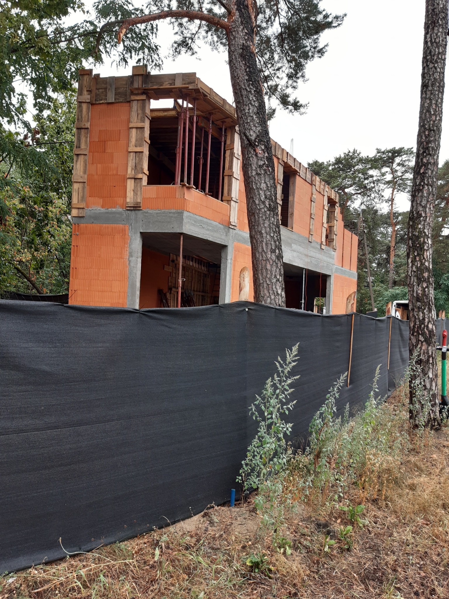 Budowa domu z czerwonej cegły, widok zza czarnej siatki ogrodzeniowej, drzewa w tle. Widoczne drewniane szalunki i stalowe podpory stropu.