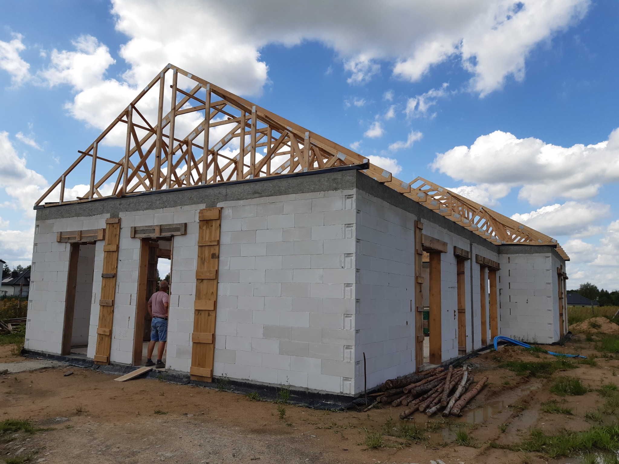 Budowa domu z białych bloczków: ściany wzniesione, drewniana konstrukcja dachu w trakcie montażu, widoczna osoba wewnątrz budynku. Niebo z chmurami w tle.