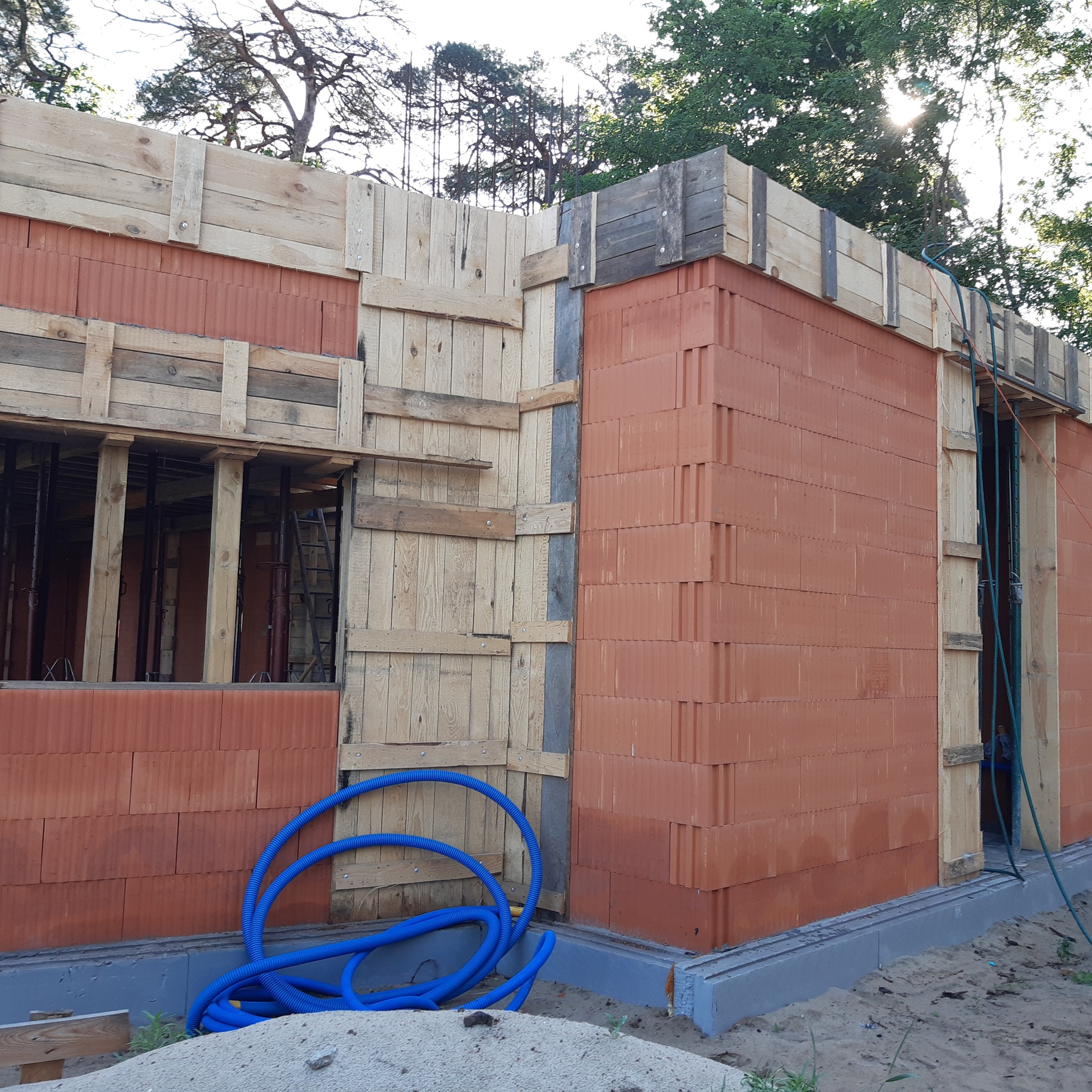 Budowa domu z pustaków ceramicznych: ściany w trakcie wznoszenia, drewniane szalunki, niebieskie rury na fundamencie. Widok z zewnątrz, słoneczny dzień.