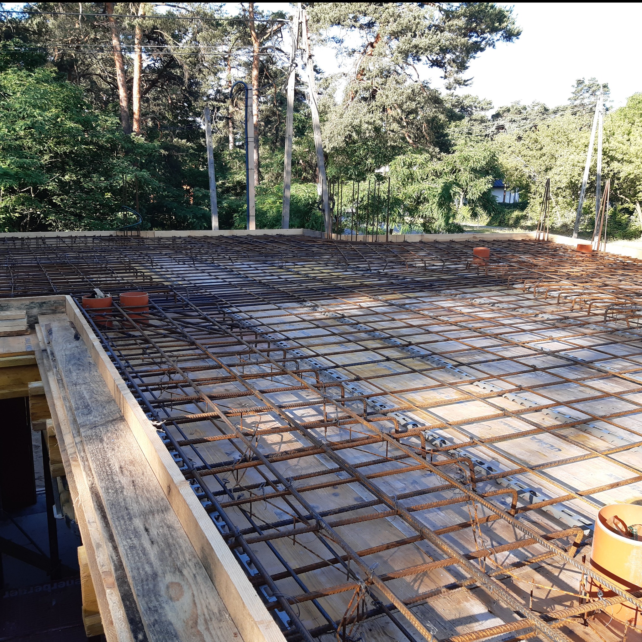 Zbrojenie płyty fundamentowej z widocznymi elementami szalunku i pionowymi rurami wentylacyjnymi na tle drzew. Etap przed betonowaniem.