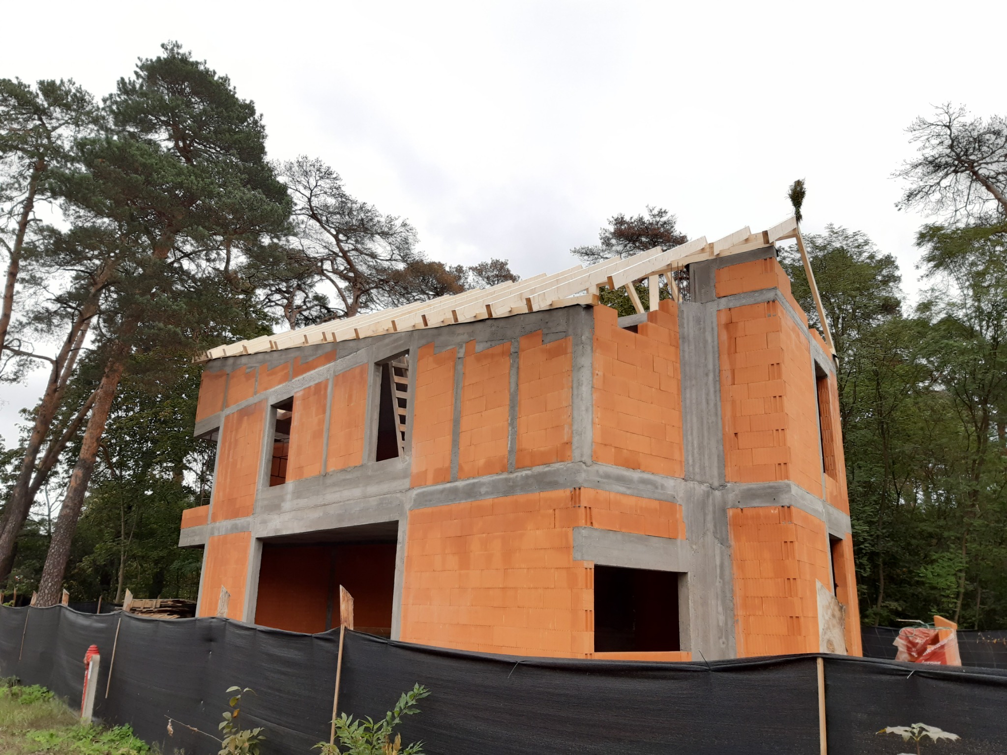 Dwukondygnacyjny dom w trakcie budowy z czerwonej cegły, z widoczną konstrukcją dachu i szarymi elementami betonowymi, otoczony zielenią i czarną siatką.
