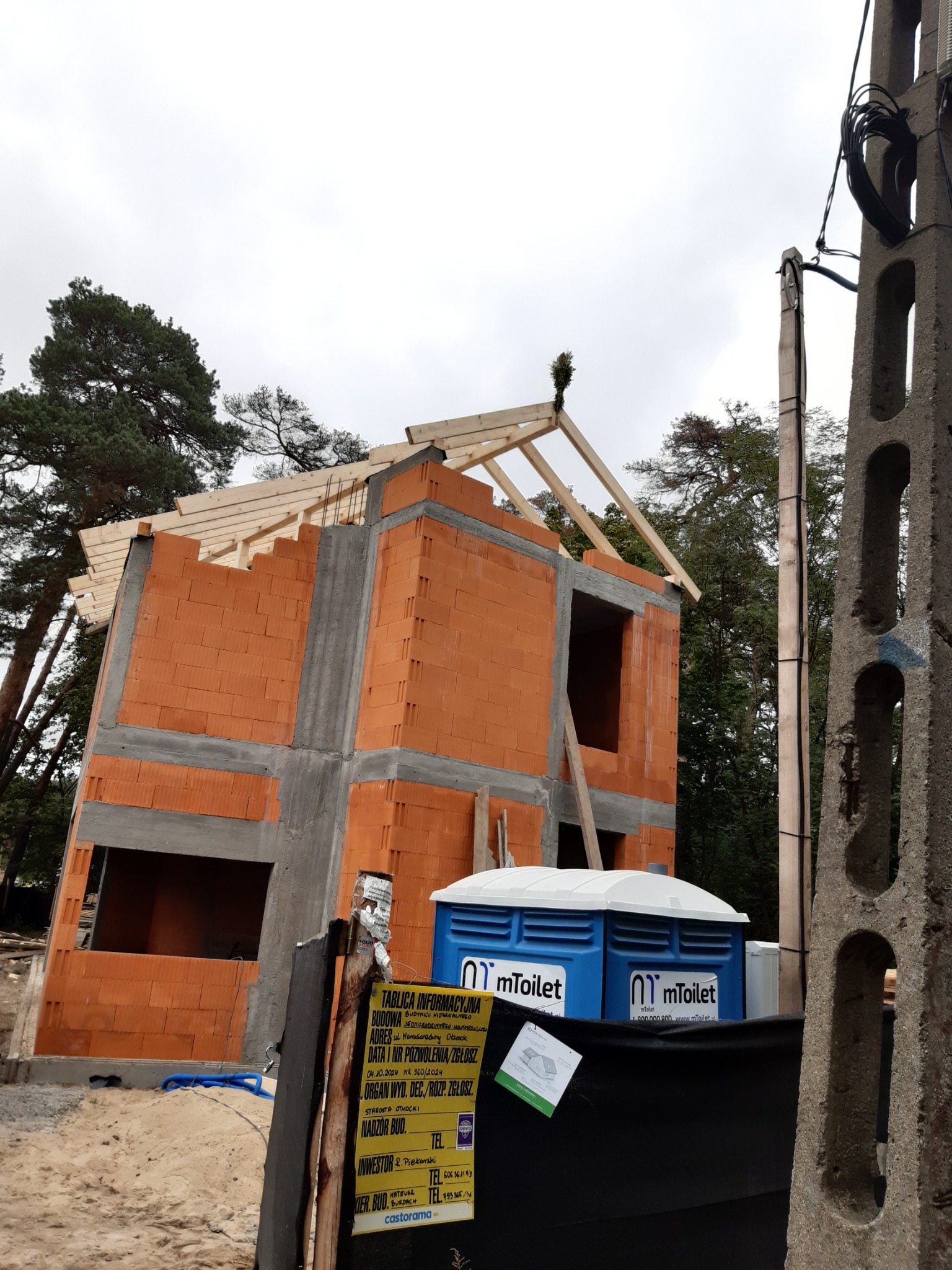 Surowy stan domu z czerwonej cegły z drewnianą konstrukcją dachu w trakcie budowy, widoczna tablica informacyjna budowy i przenośna toaleta.