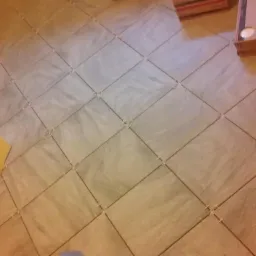 Ułożona pod kątem jasna, kwadratowa podłoga z płytek ceramicznych z krzyżykami dystansowymi. W tle kartony płytek i fragment żółtej ściany.