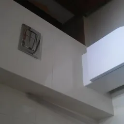 Biała toaleta podwieszana z chromowanym przyciskiem spłukującym na tle jasnych płytek w łazience. Na ścianie widoczny składany metr.