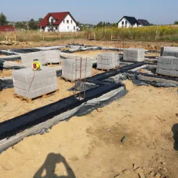 Fundamenty domu w trakcie budowy, widoczne bloczki betonowe na paletach, izolacja przeciwwilgociowa w wykopach oraz zbrojenia, w tle zabudowa jednorodzinna i błękitne niebo.