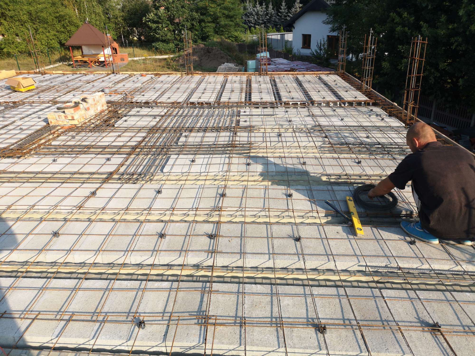 Przygotowanie zbrojenia na stropie z pustaków szalunkowych, widoczny pracownik z narzędziami, poziomica i zwój drutu wiązałkowego, w tle dom w budowie i zieleń.