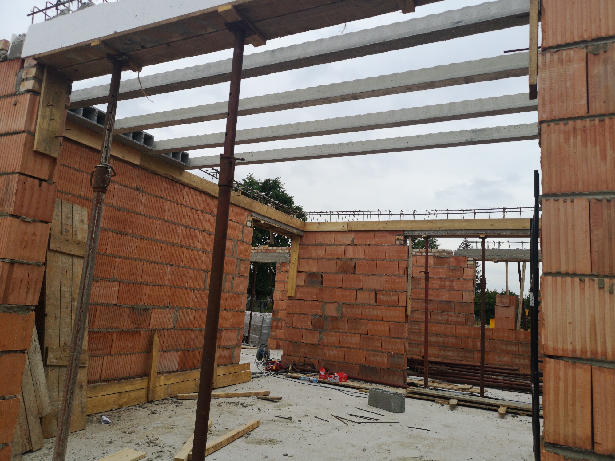 Wznoszone ściany z czerwonej cegły ceramicznej na budowie domu, widoczne betonowe nadproża i stalowe podpory szalunkowe.