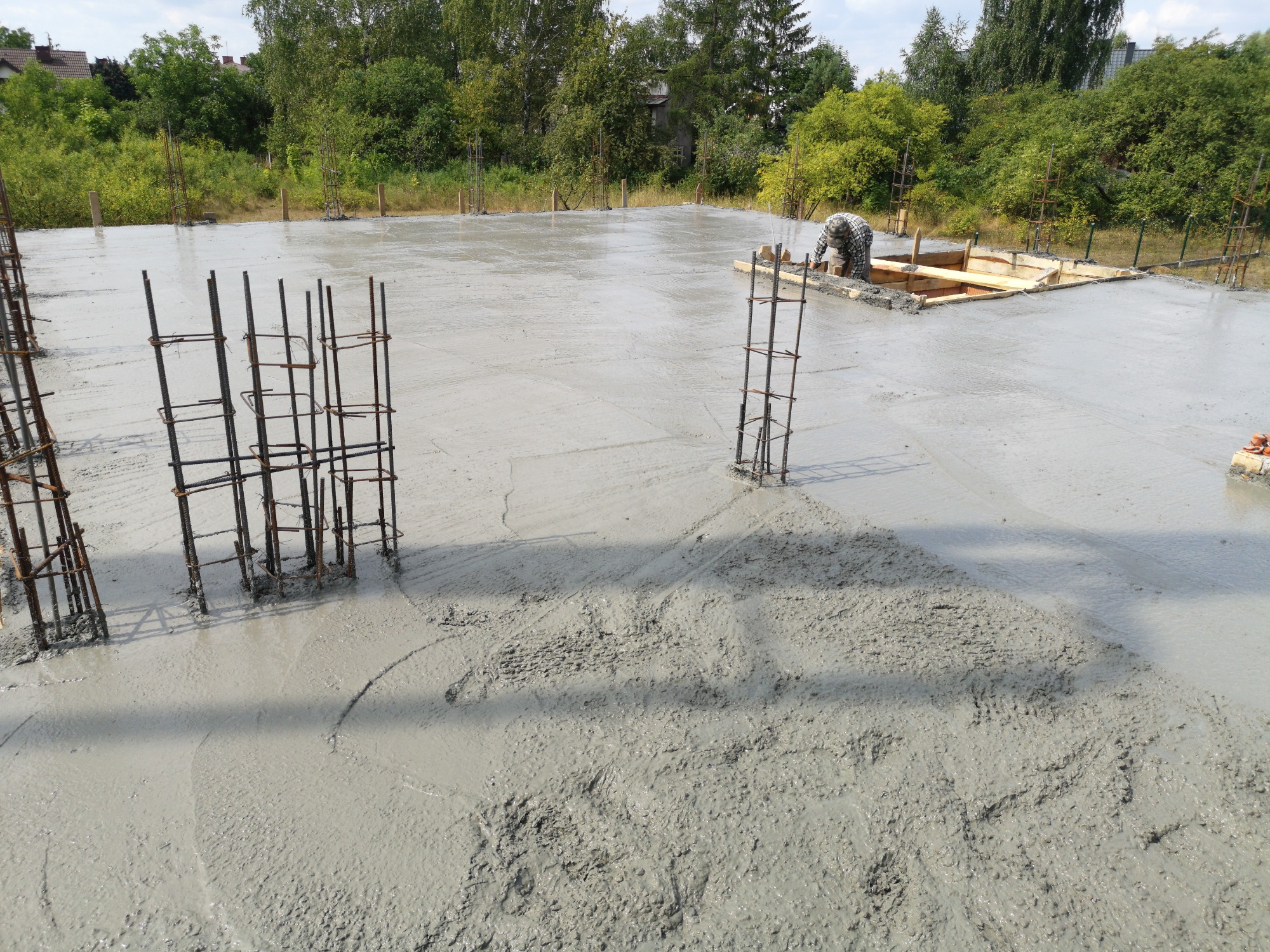Wylany beton na fundamentach z wystającymi stalowymi zbrojeniami i pracownikiem przy drewnianej szalunkowej konstrukcji w tle.