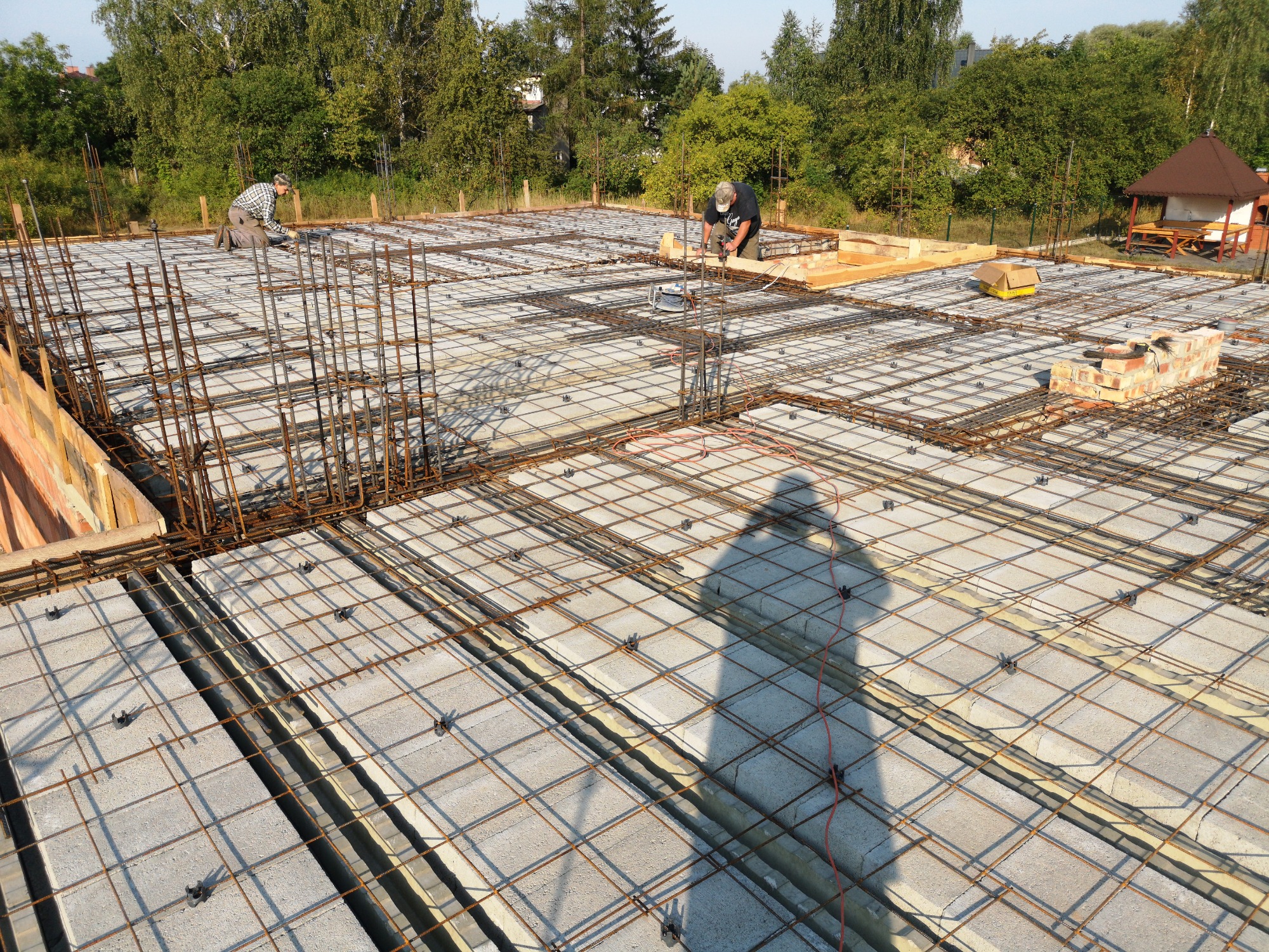 Budowa fundamentów domu w Myszkowie: widok z góry na zbrojenie i betonowe bloczki, dwóch robotników przy pracy, cień fotografa na pierwszym planie.