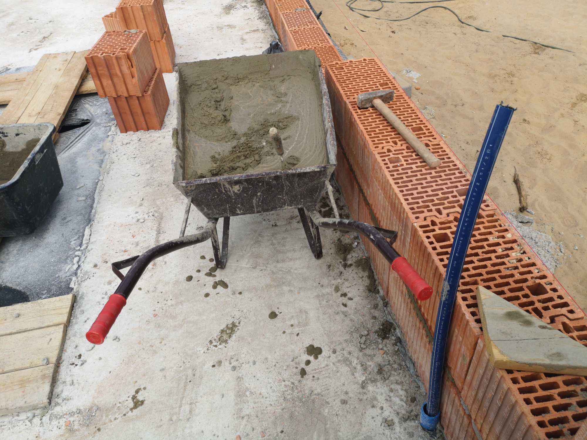 Taczka wypełniona zaprawą murarską stoi obok wznoszonej ściany z czerwonej cegły dziurawki, na której leży młotek murarski i poziomica. W tle widoczne ułożone cegły i piasek.