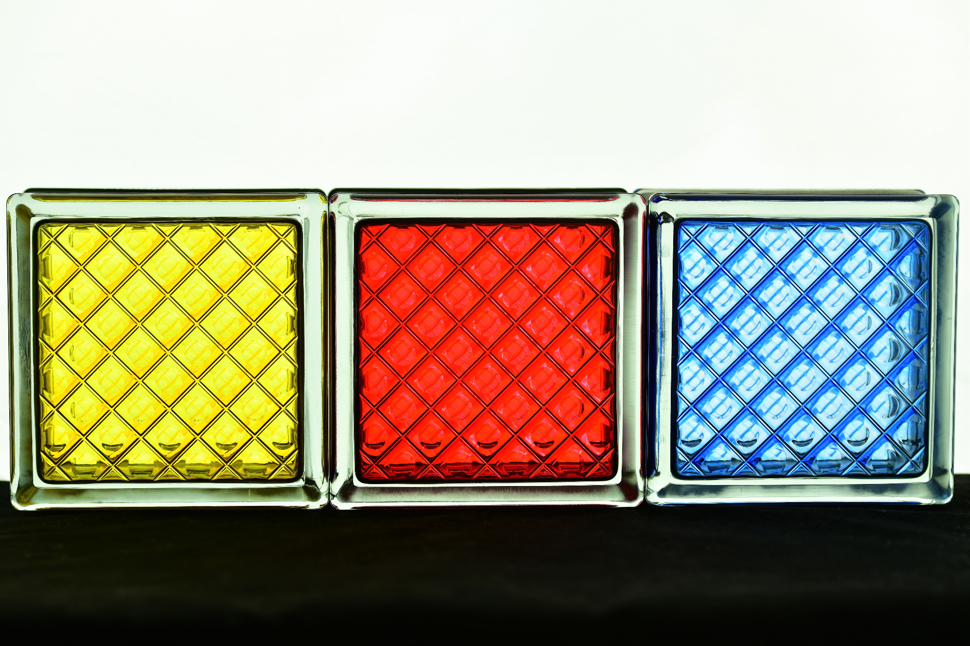 Trzy szklane pustaki w kolorach żółtym, czerwonym i niebieskim, ustawione obok siebie, tworzące dekoracyjny element architektoniczny.