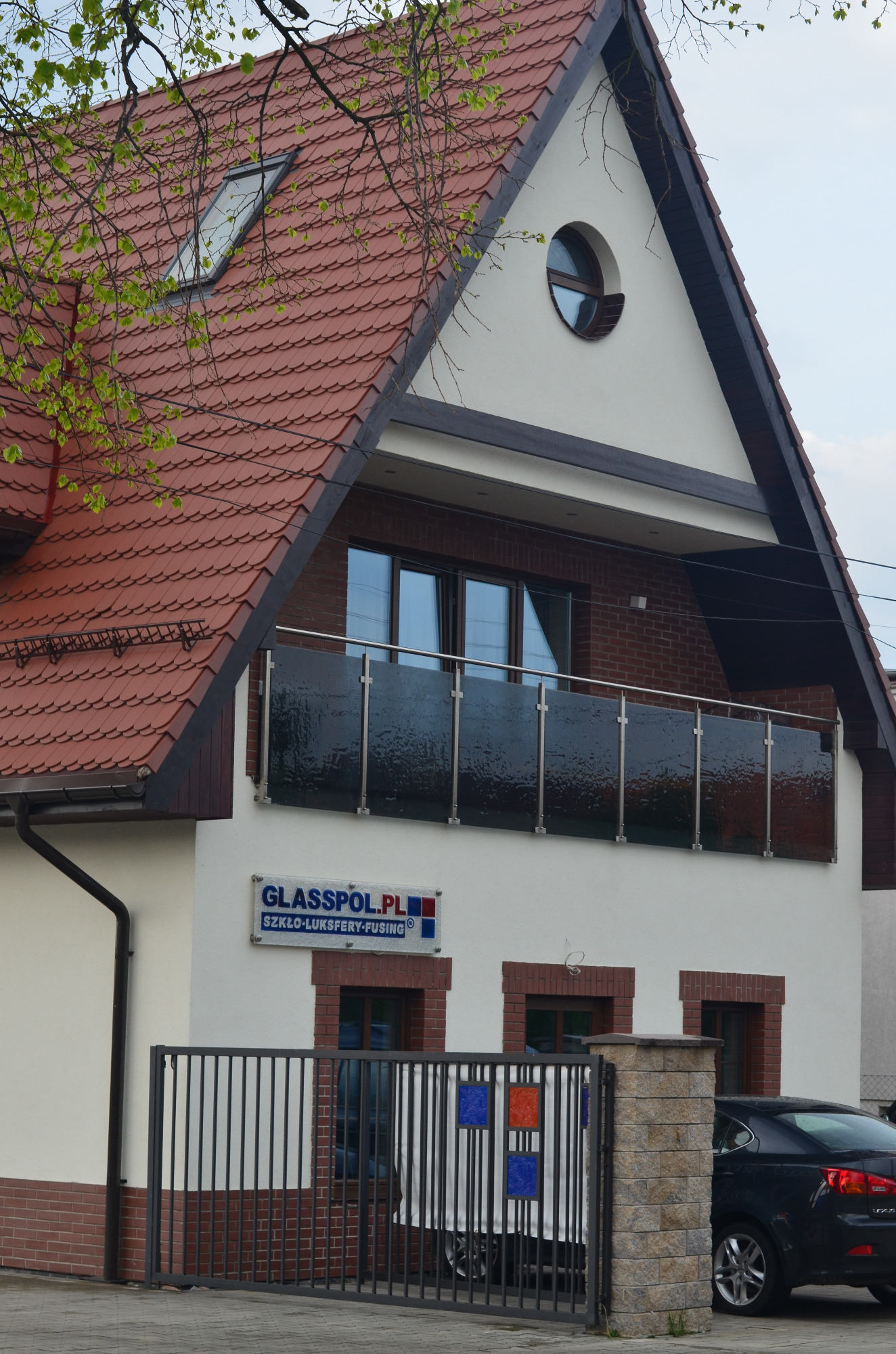 Dwupiętrowy dom z czerwonym dachem, balkonem z czarną balustradą, oknem dachowym i okrągłym oknem na szczycie, z logo firmy 'Glasspol.pl' na ścianie, widziany z poziomu ulicy przez czarny metalowy...