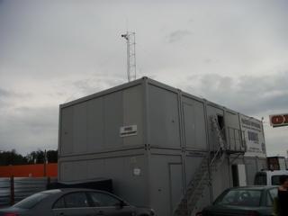 Dwukondygnacyjny, szary kontener z anteną na dachu, schodami wejściowymi i widocznymi tablicami informacyjnymi, otoczony samochodami i ogrodzeniem.