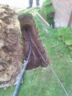 Wykop w ziemi z ułożonymi kablami teleinformatycznymi, widoczne narzędzia i fragment ściany budynku w tle.