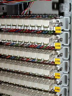 Szczegółowe ujęcie panelu krosowniczego z zamontowanymi modułami połączeniowymi, oznaczonymi numerami 10B-14B, oraz kolorowymi kablami telekomunikacyjnymi w tle, w szafie rack.