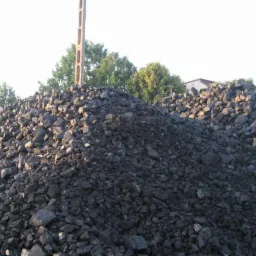 Dwa pryzmy węgla kostki na składzie, z betonowym murem w tle i słupem energetycznym po lewej stronie.