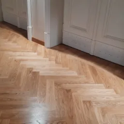 Ułożona podłoga z paneli w jodełkę, widoczne białe listwy przypodłogowe i fragmenty białych drzwi.