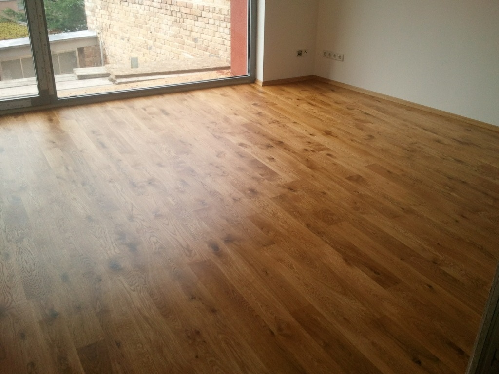 Wykończona podłoga z desek w jasnym odcieniu dębu, ułożona w nowym pokoju z dużym oknem i widokiem na ceglany budynek.