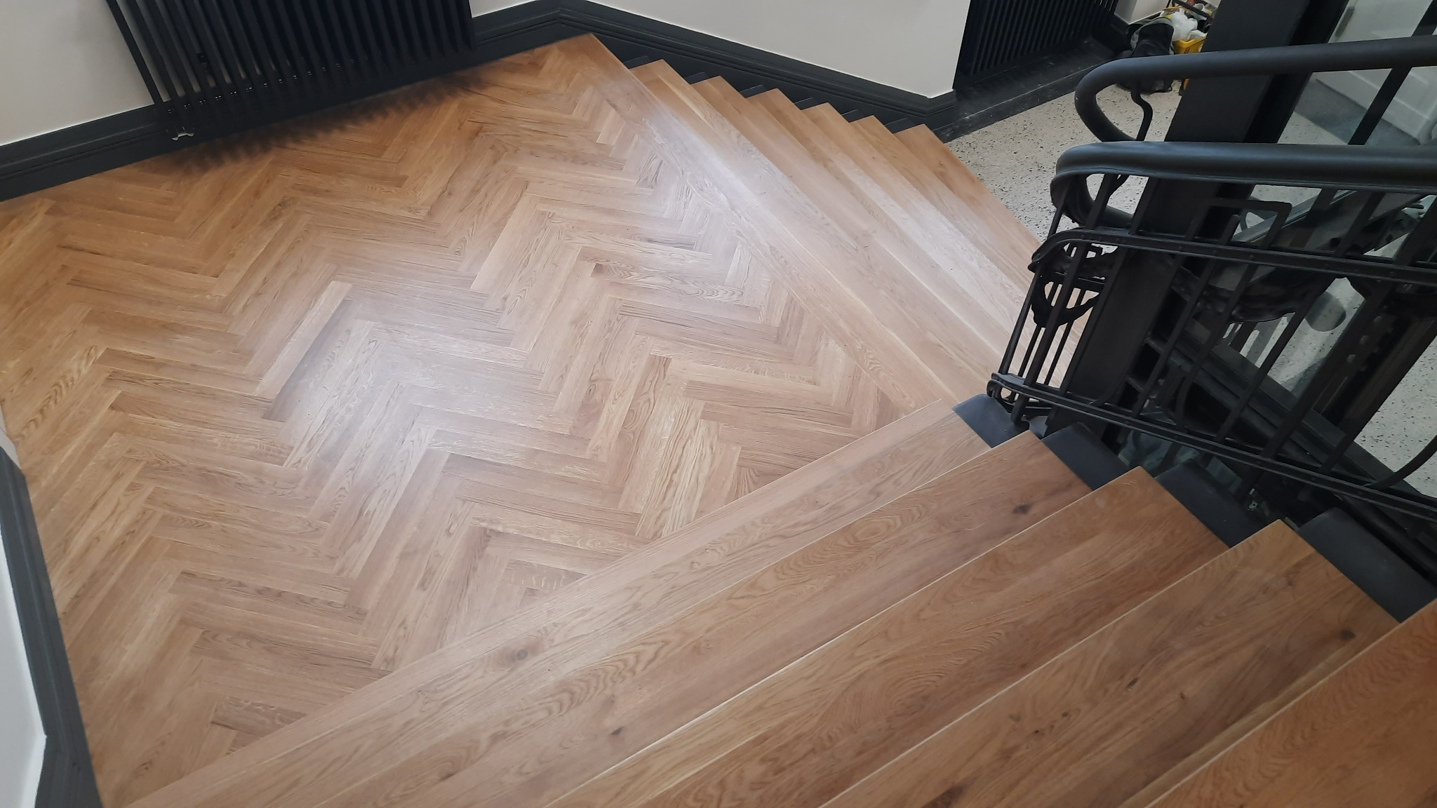 Widok z góry na schody wykończone jasnym drewnem ułożonym w jodełkę, z czarnymi metalowymi poręczami i grzejnikiem w tle.