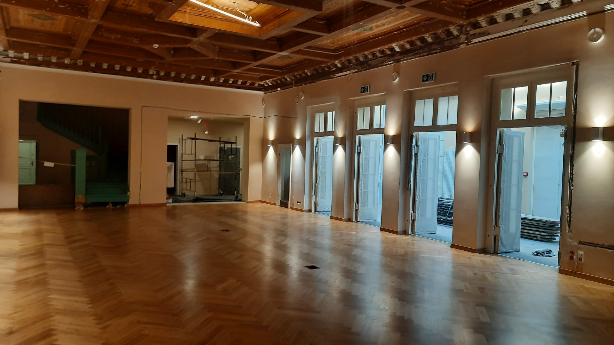 Duża sala z parkietem ułożonym w jodełkę, widoczne odnowione drewniane drzwi i sufit z dekoracyjnymi elementami, w tle rusztowania i zielone schody.