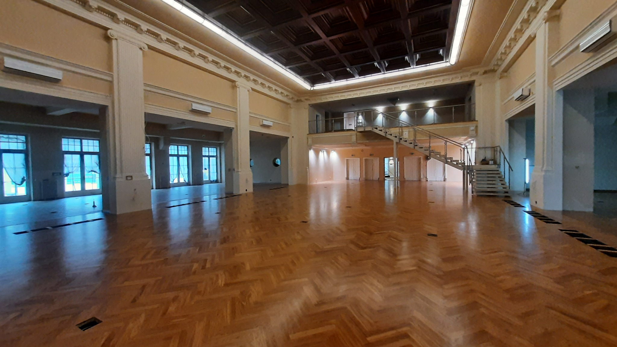 Przestronne wnętrze sali balowej z parkietem ułożonym we wzór jodełki, widoczne kolumny, antresola ze schodami i okna z zasłonami.