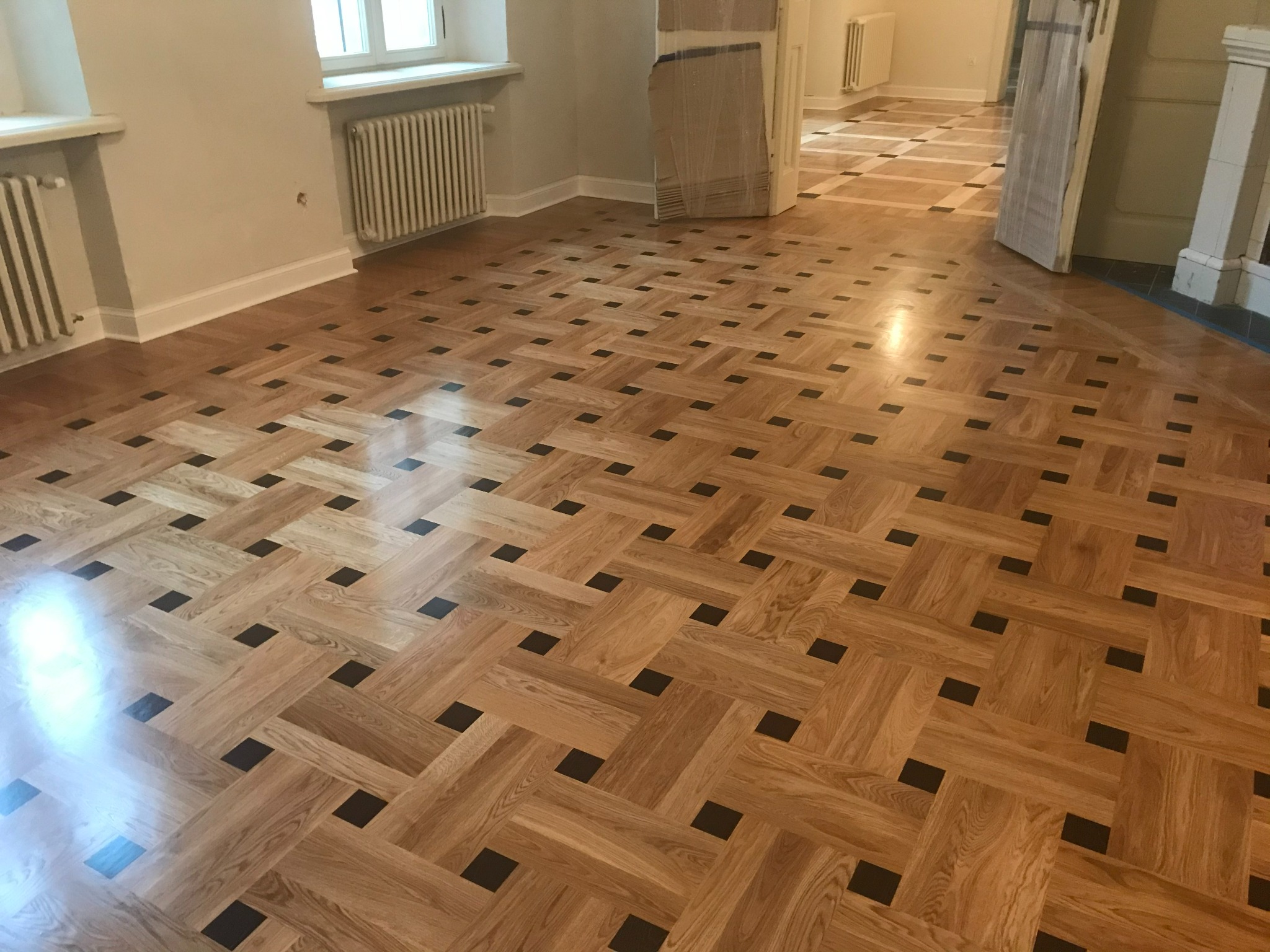 Wzór parkietu ułożonego w jodełkę z ciemnymi akcentami, widok na świeżo wykończoną podłogę w przestronnym wnętrzu z grzejnikami i częściowo zasłoniętymi drzwiami w tle.