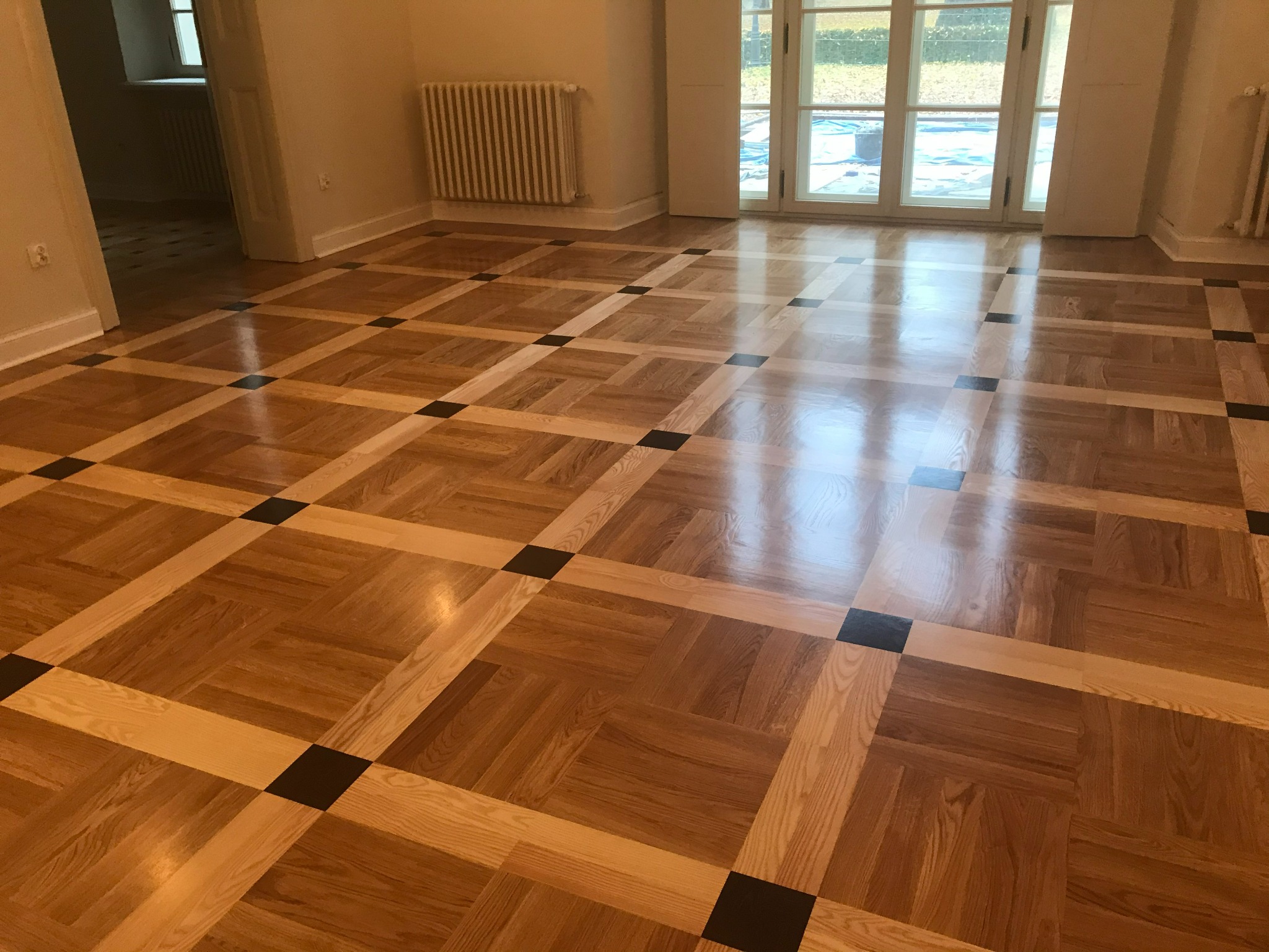 Wzór szachownicy na podłodze z polerowanego drewna, z ciemnymi kwadratami i jasnymi listwami, w przestronnym pokoju z grzejnikiem i oknami balkonowymi.