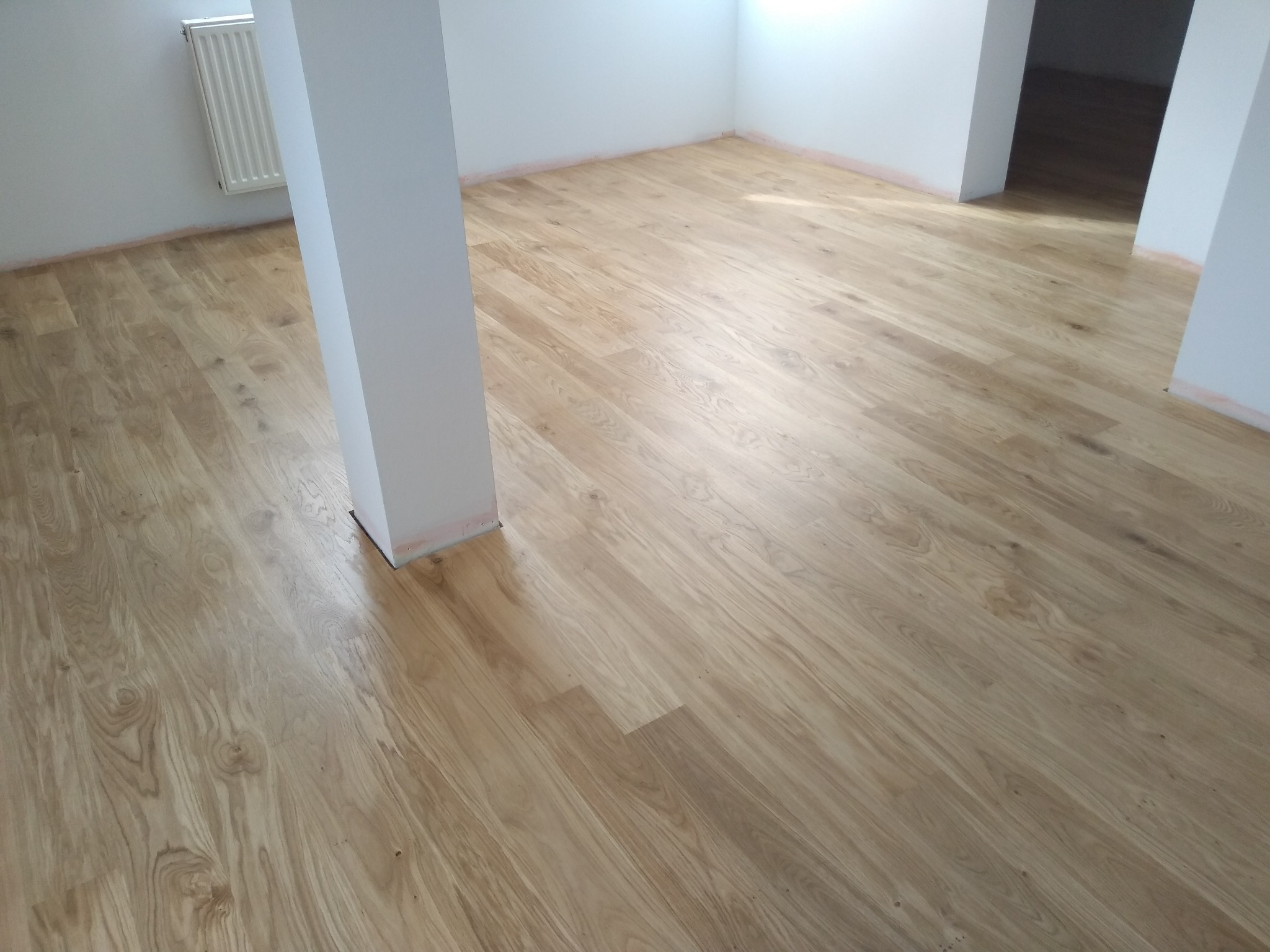 Jasna, nowo położona podłoga z desek w pustym, pomalowanym na biało pomieszczeniu z kolumną nośną, widoczne ślady po malowaniu na listwach przypodłogowych.