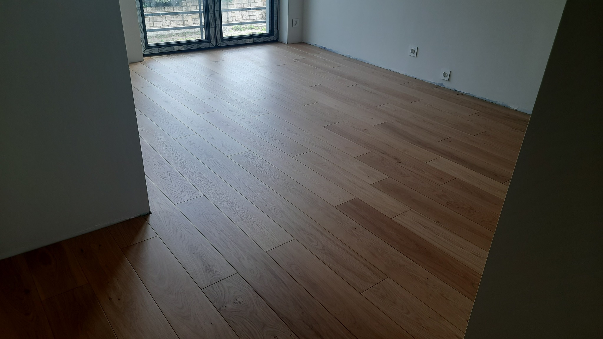 Ułożona, jasna podłoga drewniana z widocznymi słojami, w nowym wnętrzu z dużym oknem balkonowym i białymi ścianami z gniazdkami elektrycznymi.