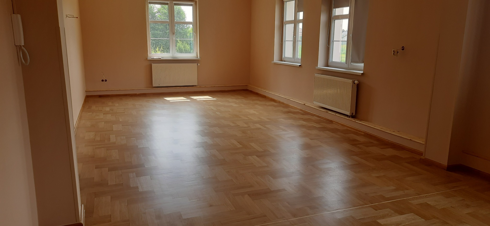 Jasny, pusty pokój z parkietem ułożonym w jodełkę, dwa okna z białymi ramami i grzejnikami pod nimi, ściany w odcieniach beżu.