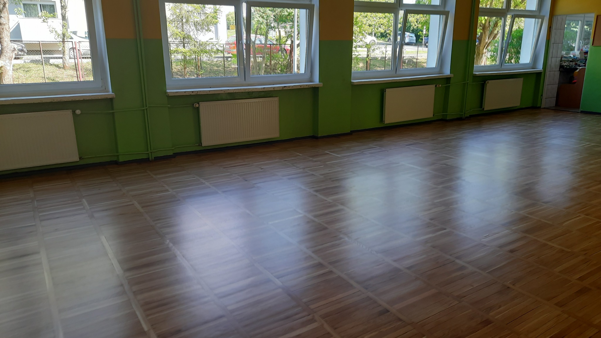 Sala z parkietem ułożonym we wzór szachownicy, ściany pomalowane na zielono, widoczne grzejniki i okna z widokiem na zieleń na zewnątrz.