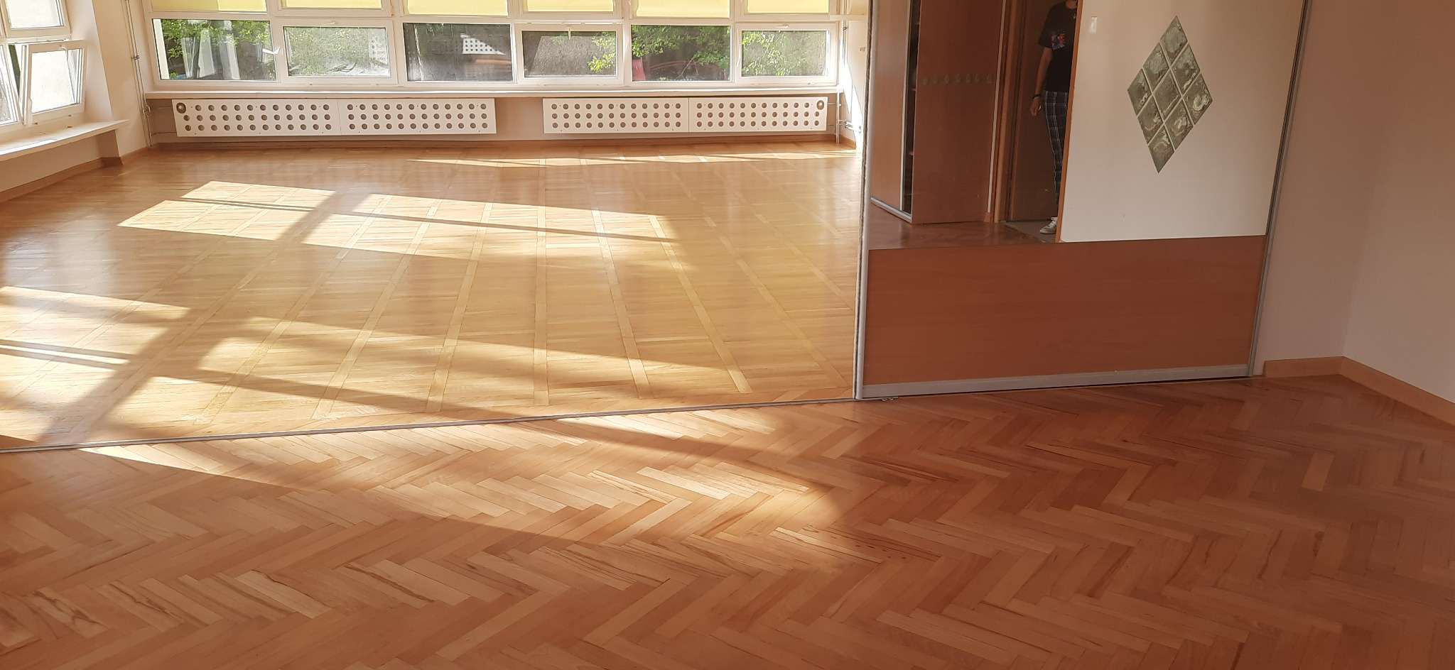 Jasna, odnowiona podłoga parkietowa ułożona we wzór jodełki, z widocznymi promieniami słonecznymi wpadającymi przez okna, w tle grzejniki i częściowo widoczne drzwi przesuwne z lustrzanym panelem.