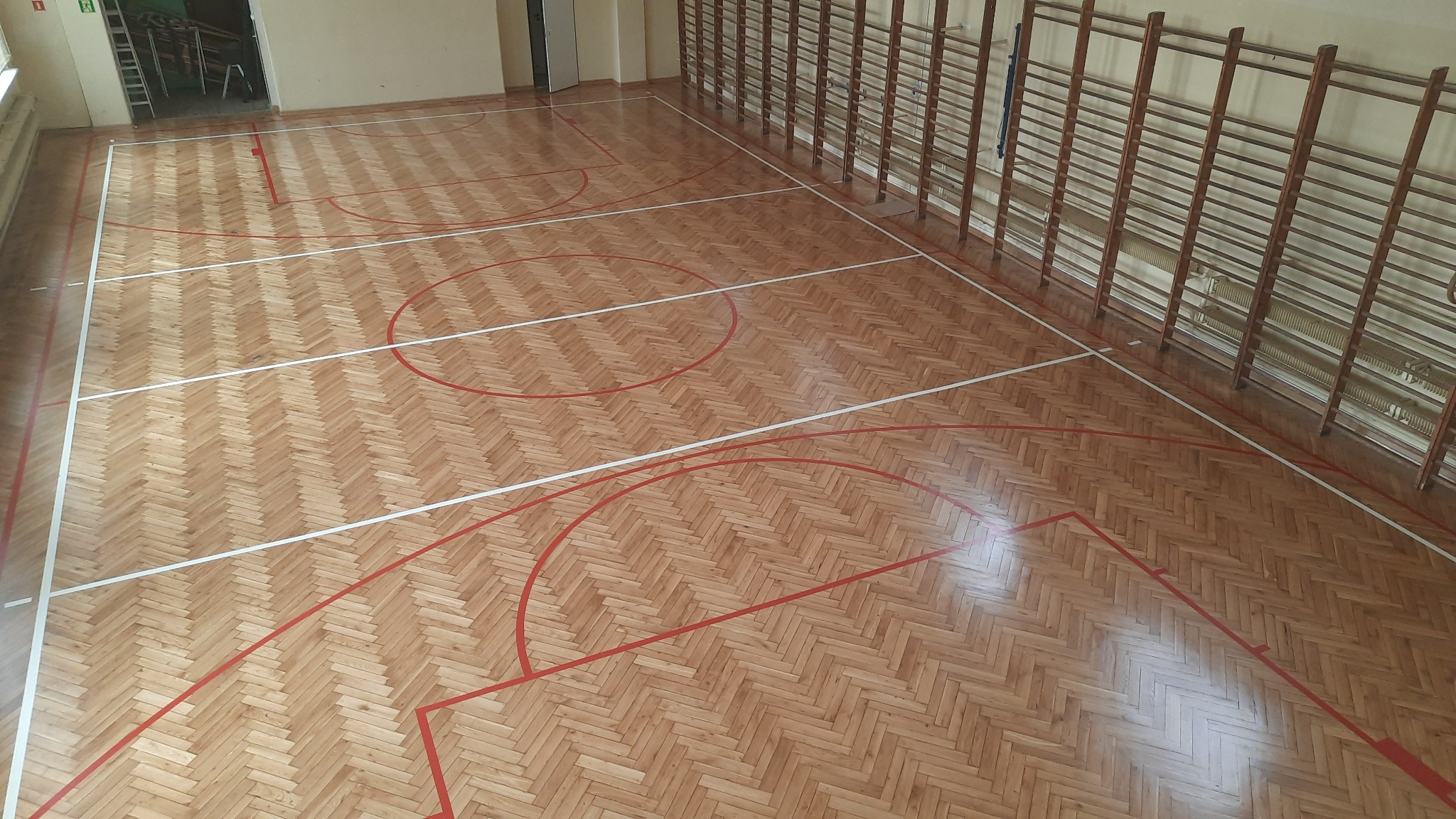 Sala gimnastyczna z parkietem ułożonym w jodełkę, pomalowanym w linie boiska do koszykówki, drabinki gimnastyczne przy ścianie.