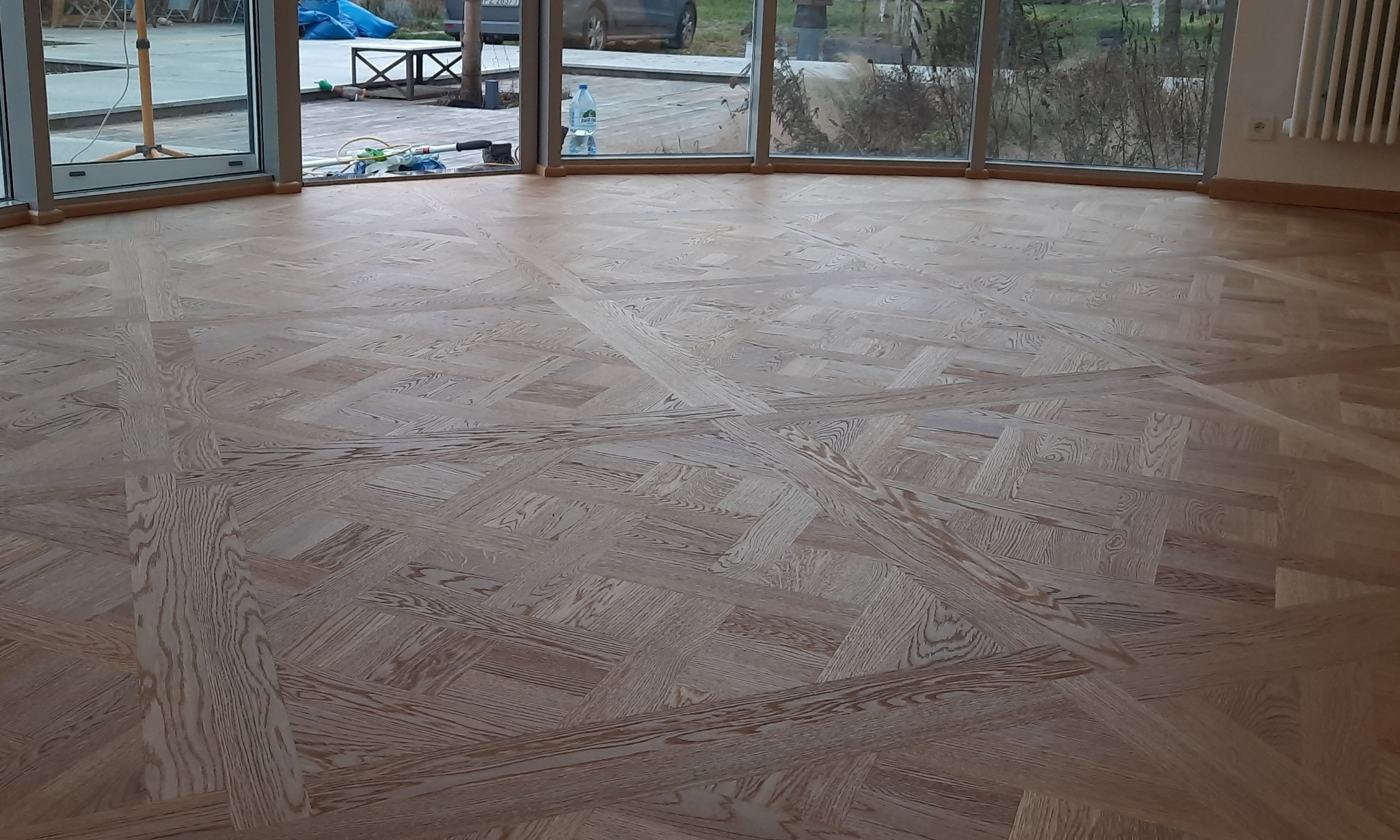 Jasna drewniana podłoga ułożona we wzór kwadratów i pasów, widok na przeszklone okna z widokiem na zewnątrz, butelka wody w tle.