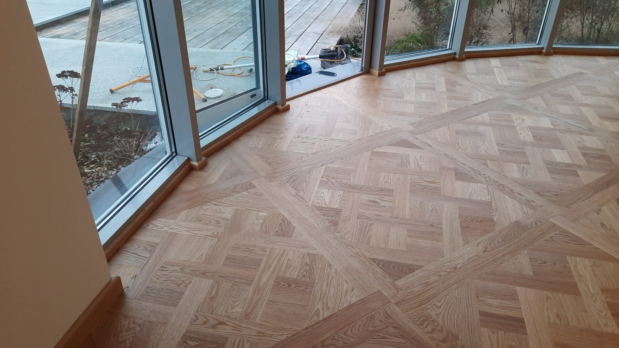 Jasna, wzorzysta podłoga ułożona z drewnianych elementów w pomieszczeniu z dużymi oknami. Widoczne narzędzia i akcesoria do montażu przy oknie.