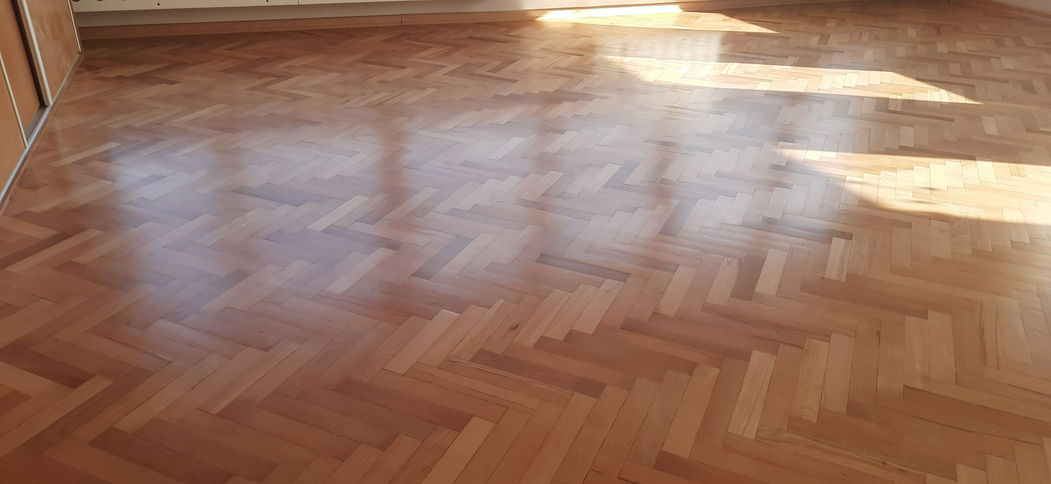 Jasna, lakierowana podłoga ułożona w jodełkę, oświetlona promieniami słońca wpadającymi przez okno.