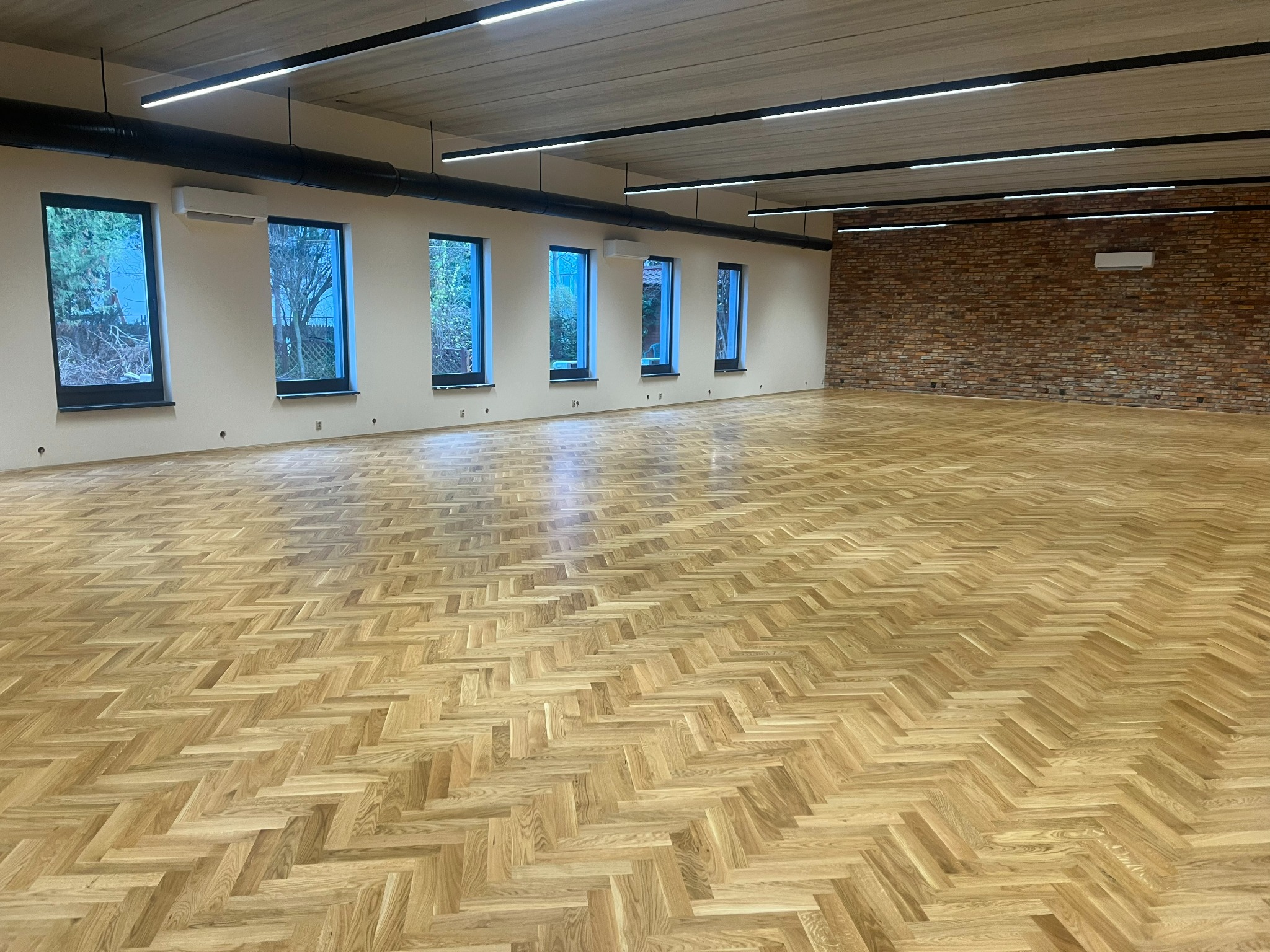 Nowoczesne wnętrze z jasną, parkietową podłogą ułożoną we wzór jodełki francuskiej, widoczne okna z widokiem na drzewa i ceglana ściana w tle.