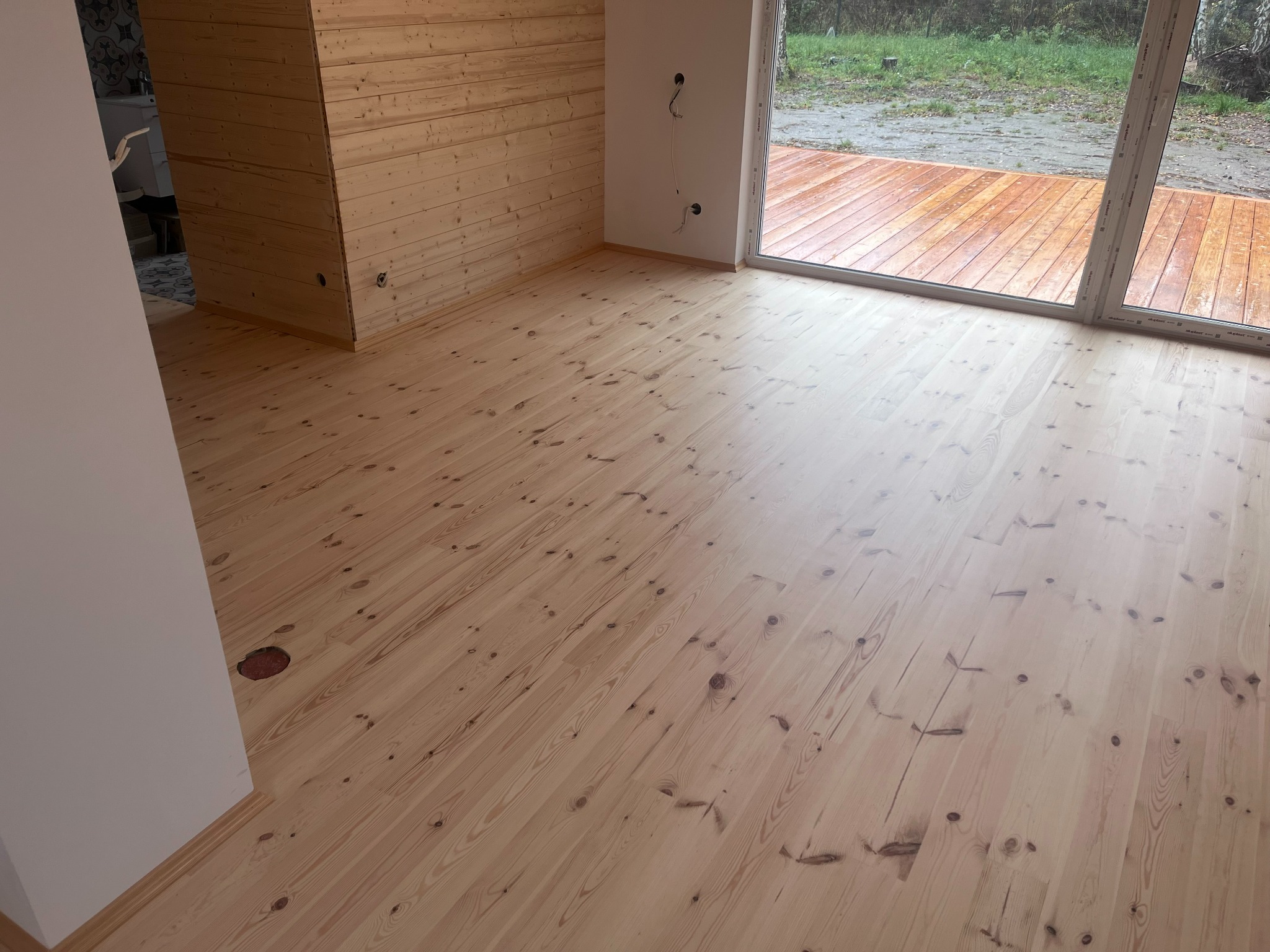 Jasna, drewniana podłoga z widocznymi słojami, świeżo ułożona w nowym wnętrzu. Ściany wykończone drewnem i bielą, duże okno z widokiem na zewnątrz.