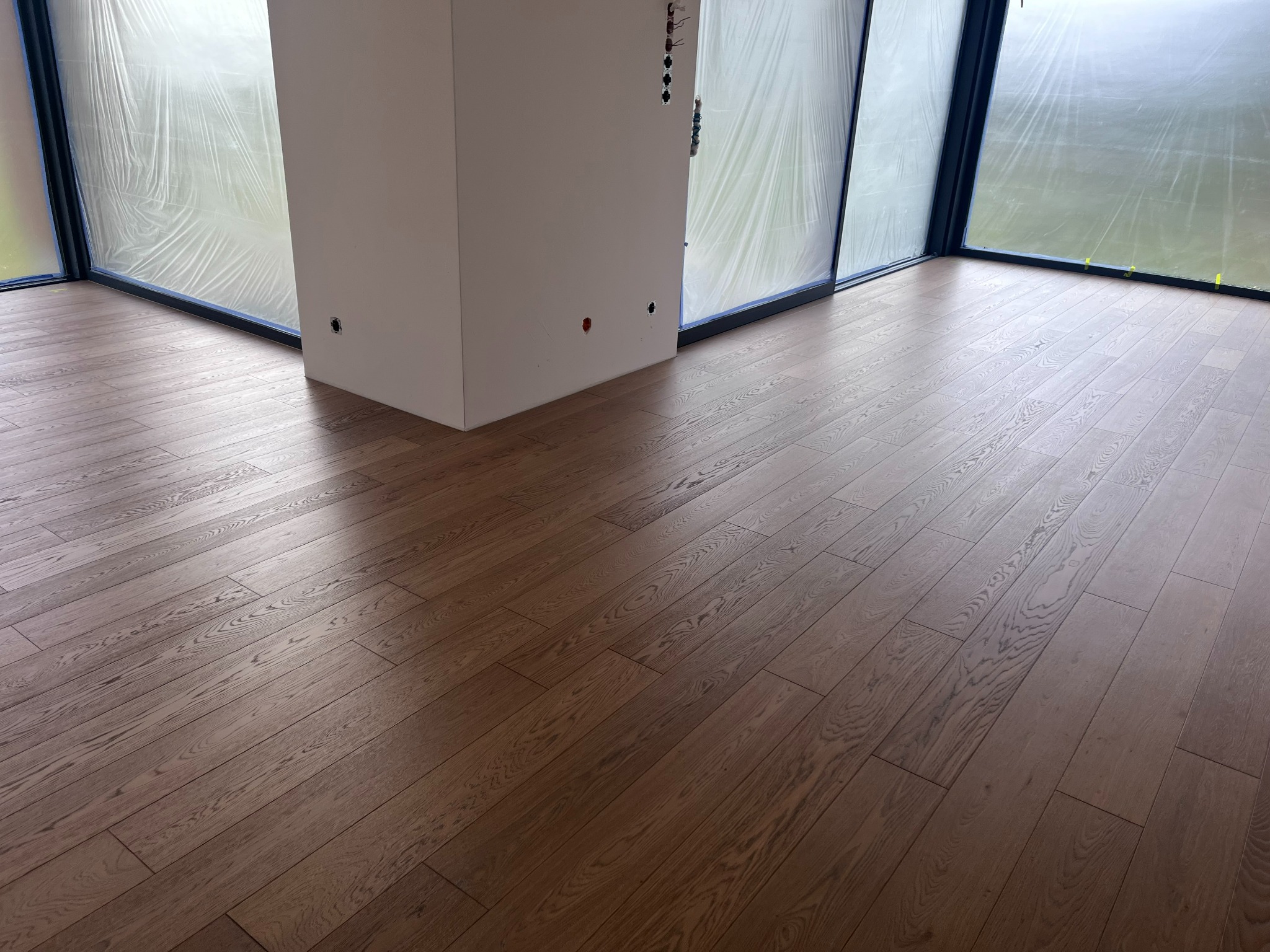 Ułożona, brązowa podłoga z desek w nowym wnętrzu. Okna zabezpieczone folią malarską, widoczne gniazdka elektryczne w ścianie. Jasne, naturalne światło.