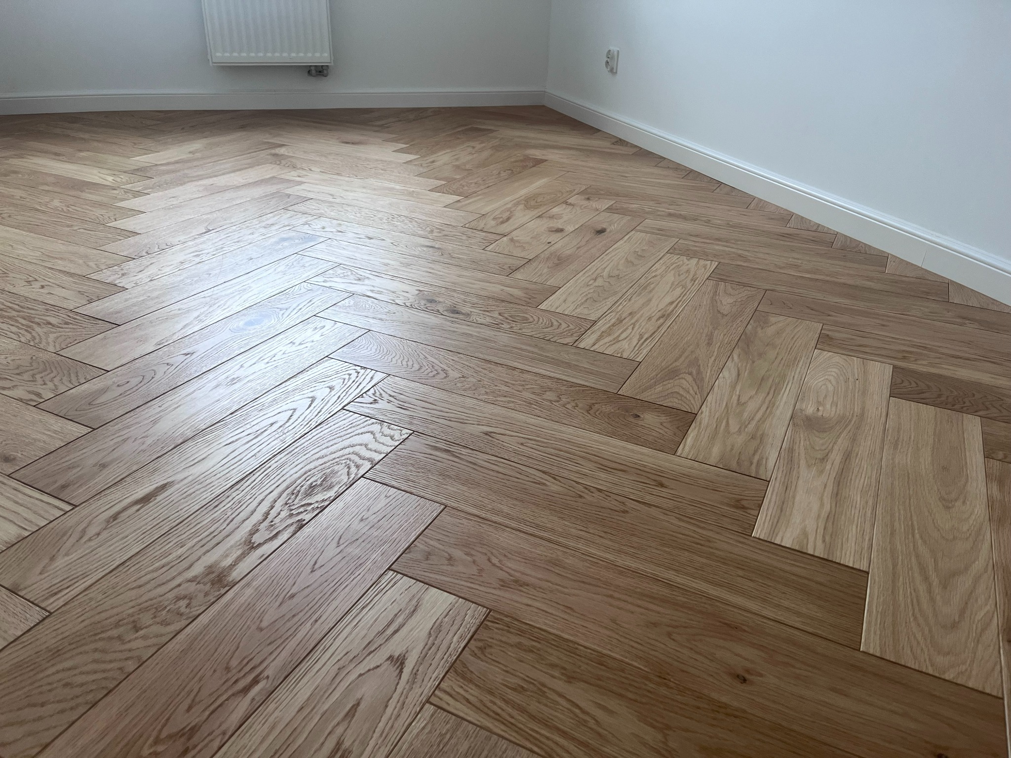 Jasna, dębowa podłoga ułożona we wzór jodełki francuskiej, z widoczną listwą przypodłogową i fragmentem grzejnika na białej ścianie. Naturalne oświetlenie.