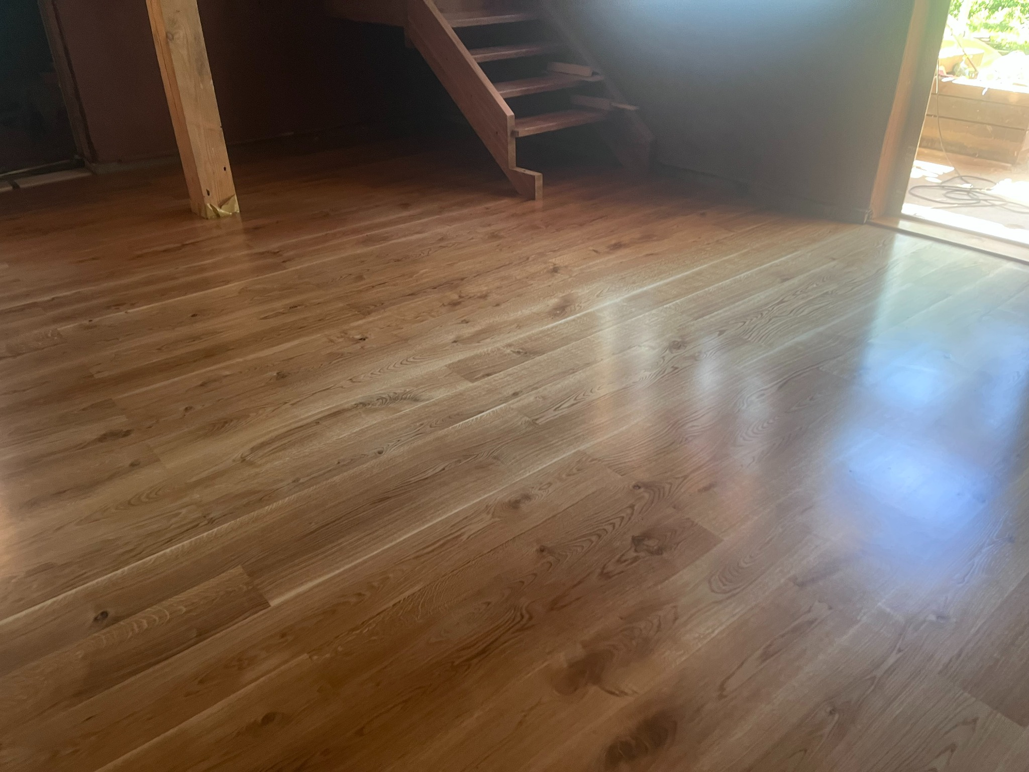 Jasna, drewniana podłoga ułożona pod schodami, z widocznym słupem konstrukcyjnym zabezpieczonym taśmą. Naturalne światło podkreśla strukturę drewna.