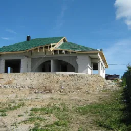 Niedokończony dom z zielonym dachem i białymi ścianami z betonu komórkowego, widoczny z poziomu gruntu. W tle błękitne niebo i fragment ogrodzenia z siatki.