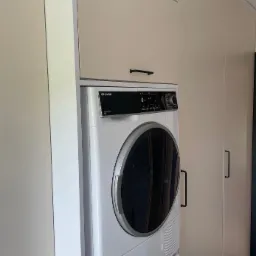 Pralka i suszarka w zabudowie meblowej z beżowymi frontami i czarnymi uchwytami, widoczne na tle szafy i czarnej podłogi.