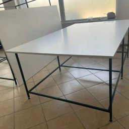 REBEL Sławomir Olek - Biały stół z czarnymi nogami ustawiony na kafelkach, z białym panelem za nim, w tle widoczne okno i elementy wyposażenia.