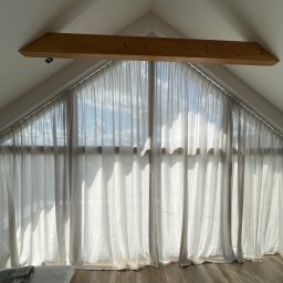 REBEL Sławomir Olek - Jasne, przesuwne zasłony w oknie o nietypowym, trójkątnym kształcie, umieszczonym na poddaszu z drewnianą belką nad oknem. Widoczne promienie słoneczne.