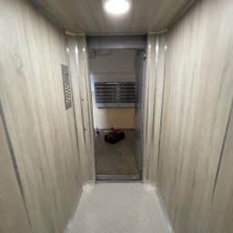 REBEL Sławomir Olek - Wnętrze windy osobowej z panelami w kolorze jasnego drewna i metalowym panelem sterowania. Widok na otwarte drzwi i korytarz z kratką wentylacyjną.