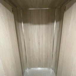 REBEL Sławomir Olek - Wnętrze windy osobowej z panelami imitującymi drewno i metalowymi listwami wykończeniowymi. Widok z dołu kabiny, neutralna kolorystyka, symetryczny układ.