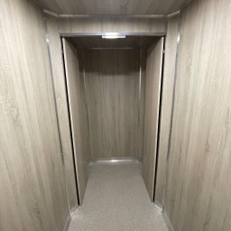 REBEL Sławomir Olek - Wnętrze windy towarowej z panelami imitującymi drewno i szarą podłogą. Metalowe wykończenia krawędzi. Widok z perspektywy osoby wchodzącej.