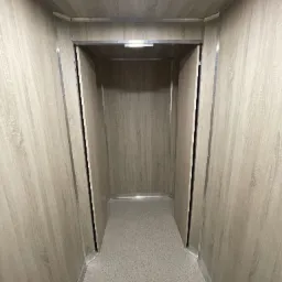 Wnętrze windy towarowej z panelami imitującymi drewno i szarą podłogą. Metalowe wykończenia krawędzi. Widok z perspektywy osoby wchodzącej.